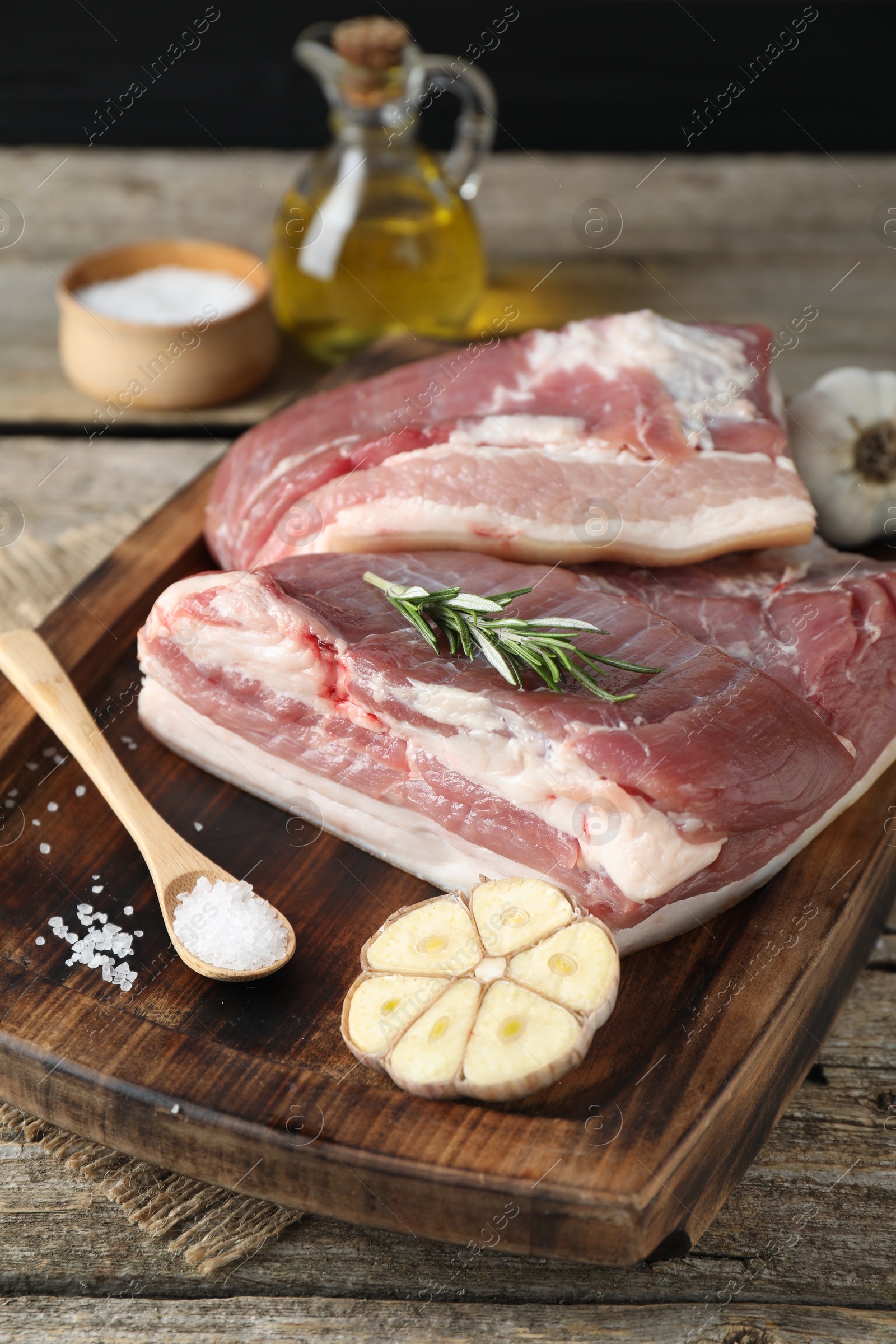 Photo of Pieces of raw pork belly, salt, garlic and rosemary on wooden table, closeup