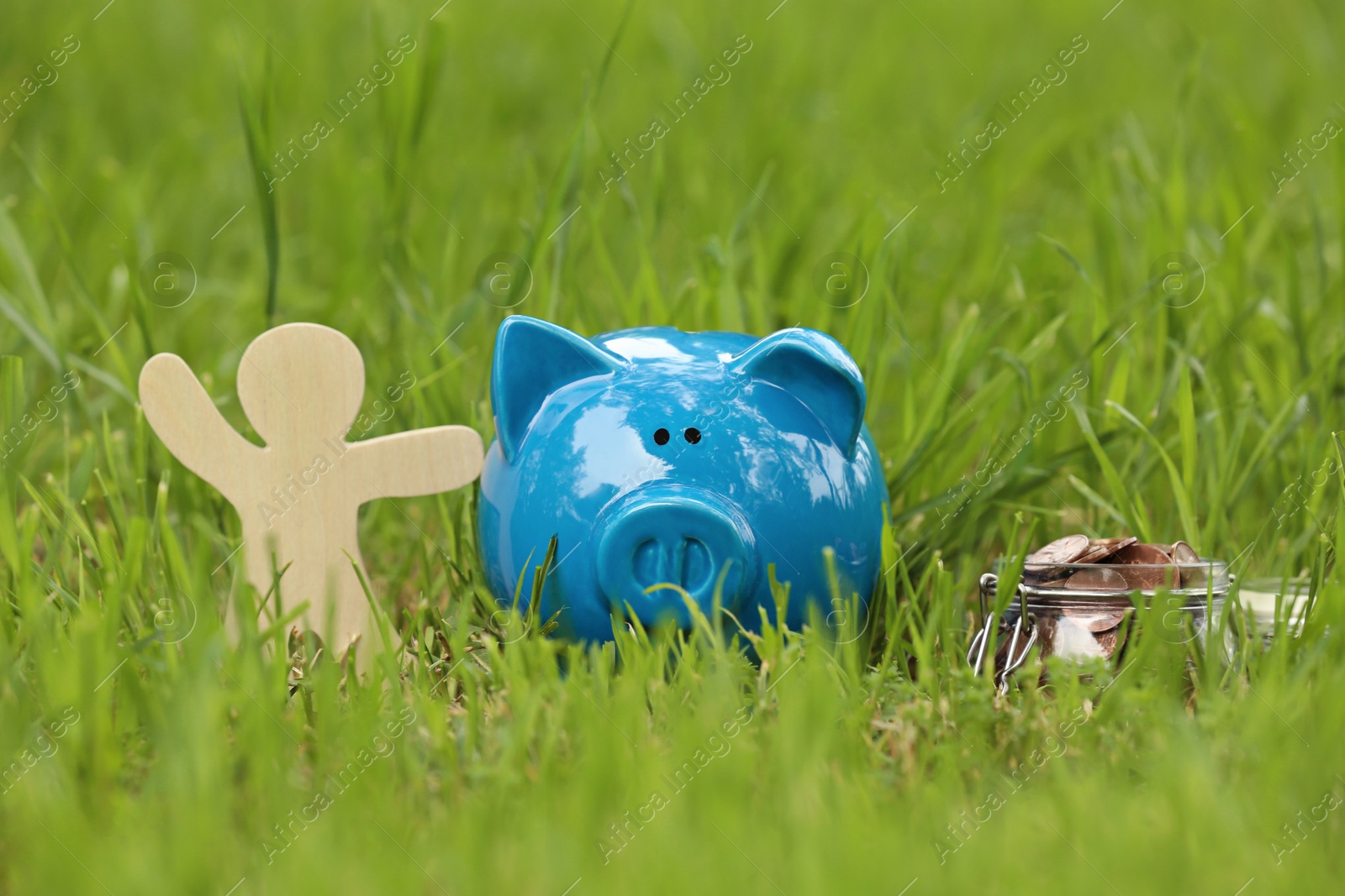 Photo of Cute piggy bank, wooden person and jar with coins on green grass in park