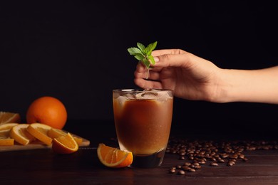 Woman putting mint into glass of refreshing drink with coffee and orange juice at wooden table, closeup