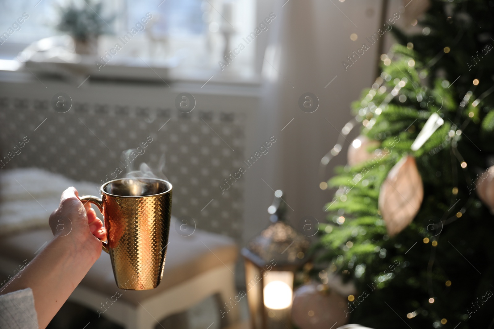 Photo of Woman with golden cup of hot drink near Christmas tree at home, closeup