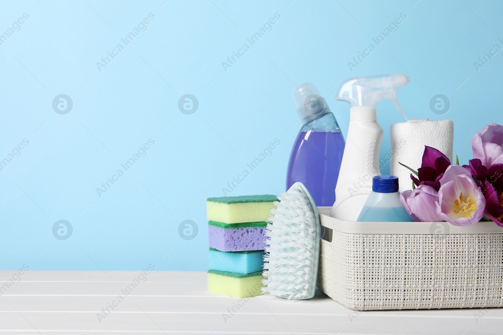 Photo of Spring cleaning. Basket with detergents, flowers and tools on white wooden table against light blue background. Space for text