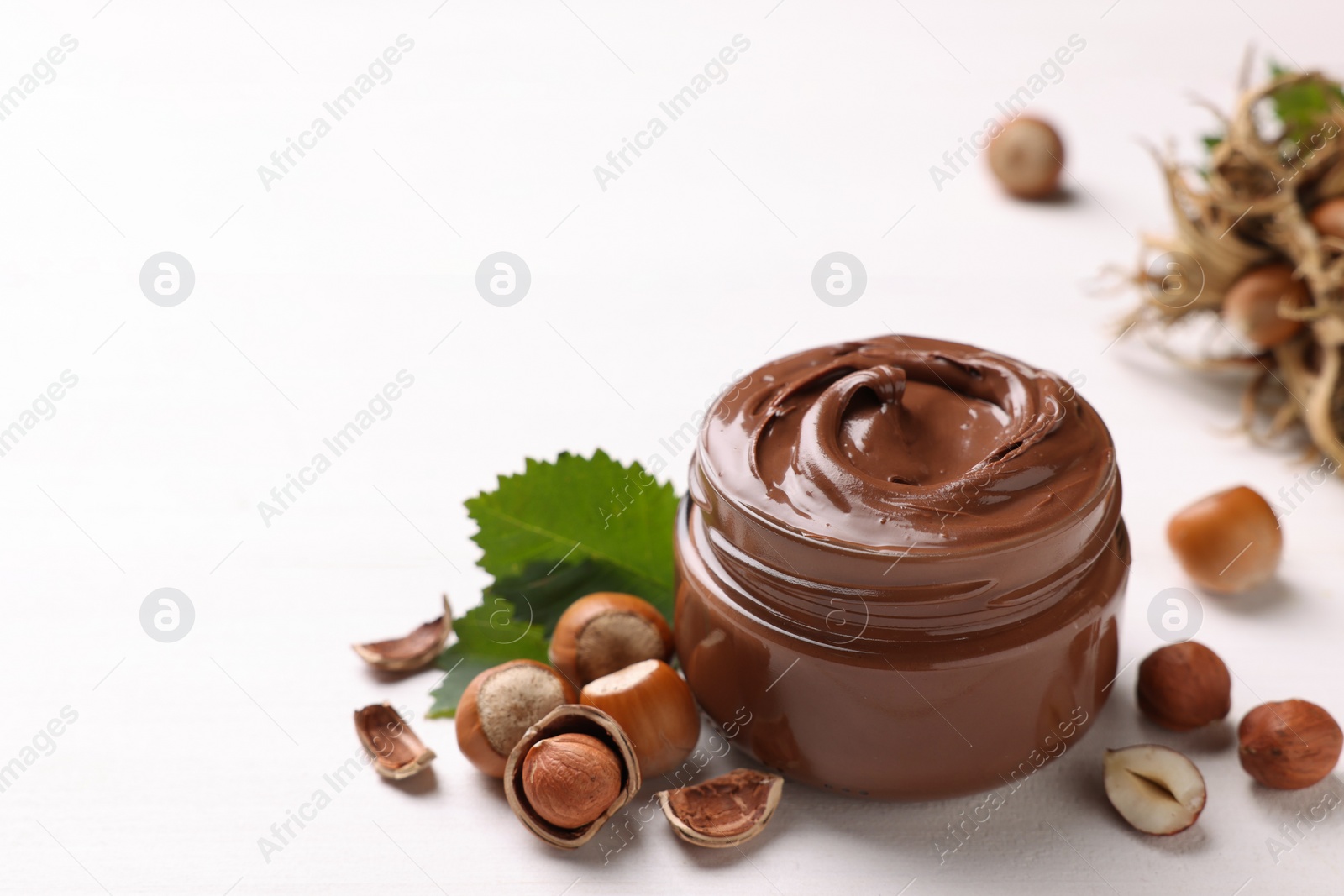 Photo of Glass jar with tasty chocolate spread, hazelnuts and green leaves on light background