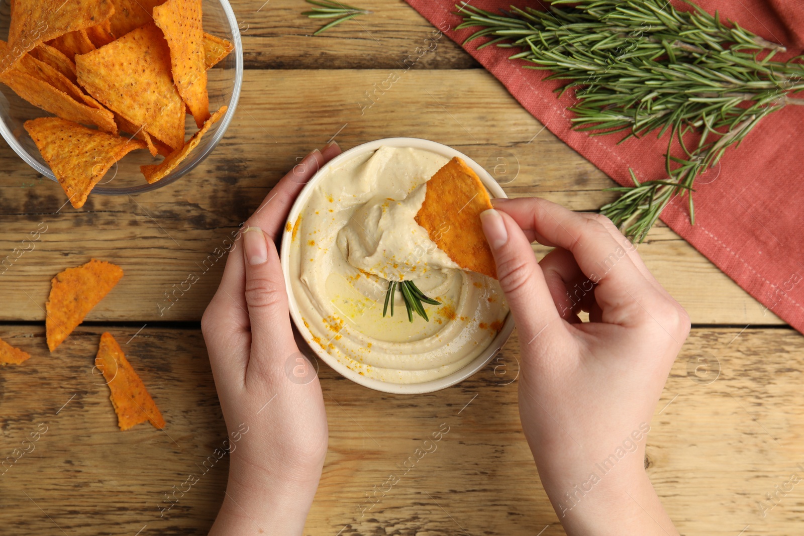 Photo of Woman dipping nacho chip into hummus at wooden table, top view