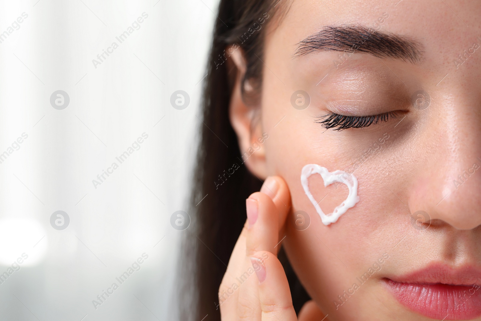 Photo of Young woman with dry skin and heart made of cream on her face indoors, closeup. Space for text