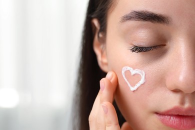 Young woman with dry skin and heart made of cream on her face indoors, closeup. Space for text