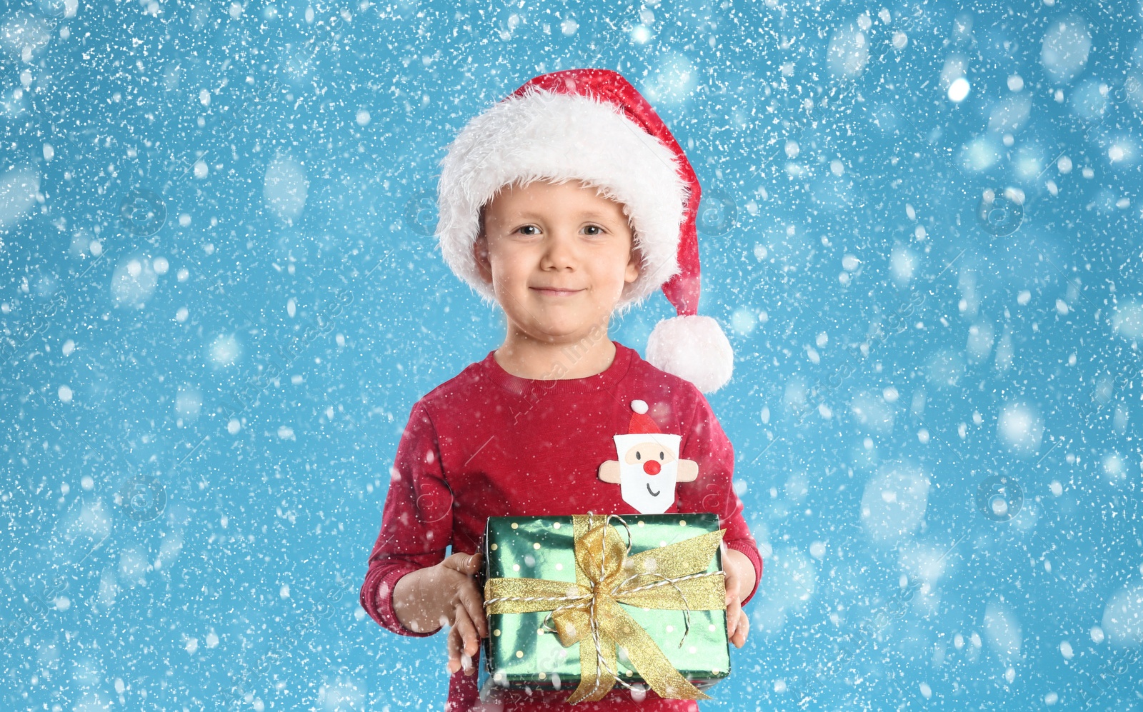 Image of Cute child in Santa hat with Christmas gift under snowfall on light blue background