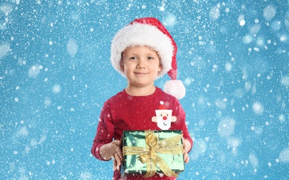 Cute child in Santa hat with Christmas gift under snowfall on light blue background