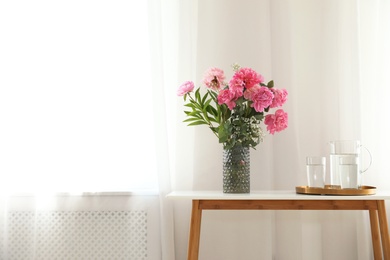 Photo of Peonies in vase and tray with jug and glasses of water on console table near wall, space for text. Room interior