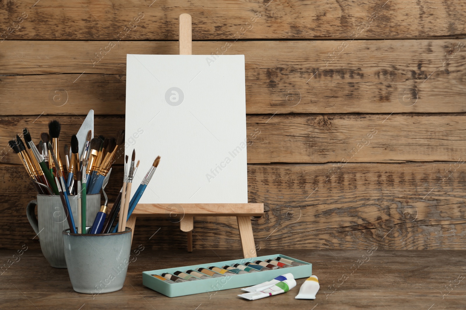 Photo of Easel with blank canvas, paints and brushes on wooden table. Space for text