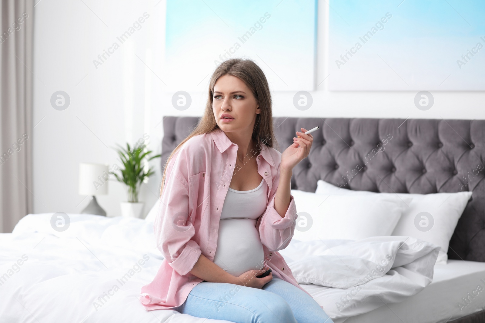 Photo of Young pregnant woman smoking cigarette in bedroom. Harm to unborn baby