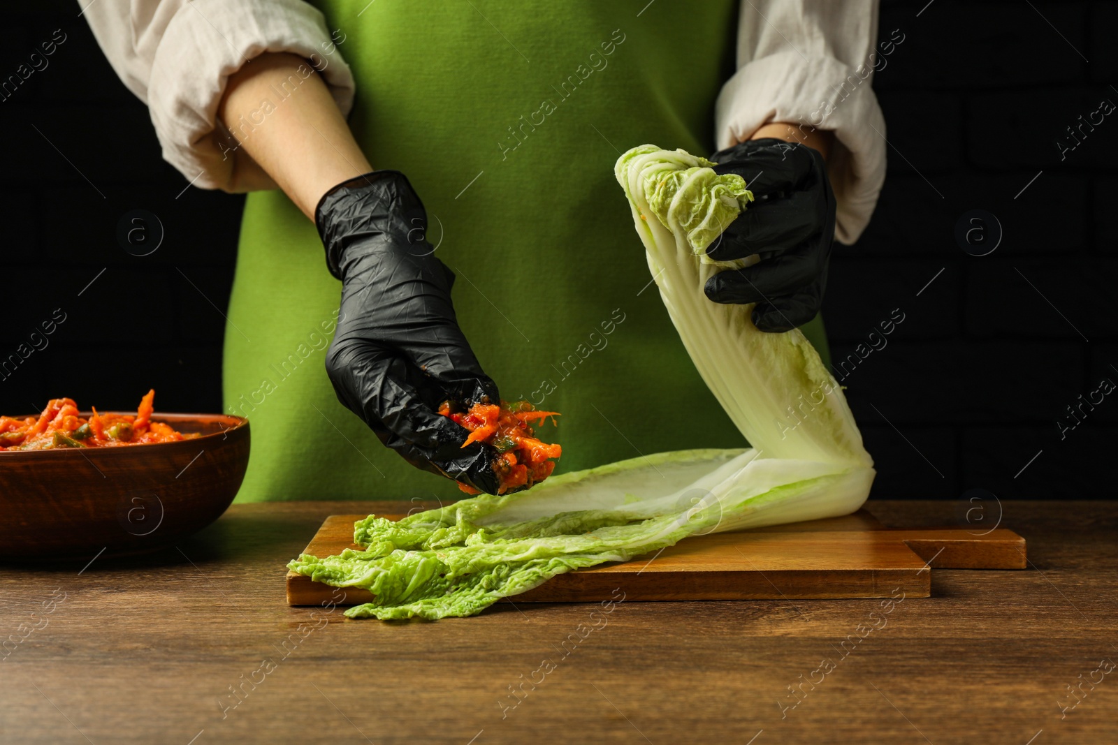 Photo of Woman preparing spicy cabbage kimchi at wooden table against dark background, closeup