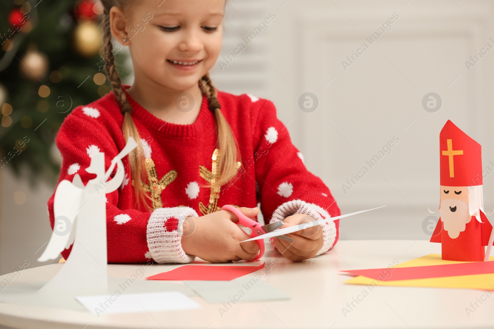 Photo of Cute little girl cutting paper at table with Saint Nicholas toy indoors