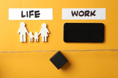 Photo of Family figures, smartphone and black cube on yellow wooden table, flat lay. Life work balance concept