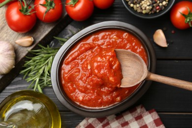 Photo of Homemade tomato sauce in bowl, spoon and fresh ingredients on black wooden table, flat lay