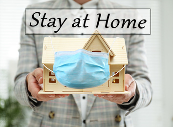 Image of Stay at home during coronavirus quarantine. Woman holding wooden house model with medical mask