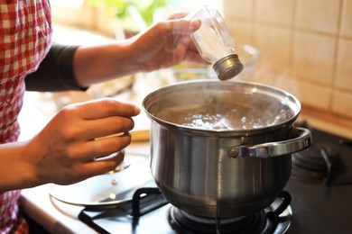 Photo of Woman adding salt to boiling water in pot on stove indoors, closeup