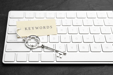 Photo of Keyboard, vintage key and tag with word KEYWORDS on black table, closeup