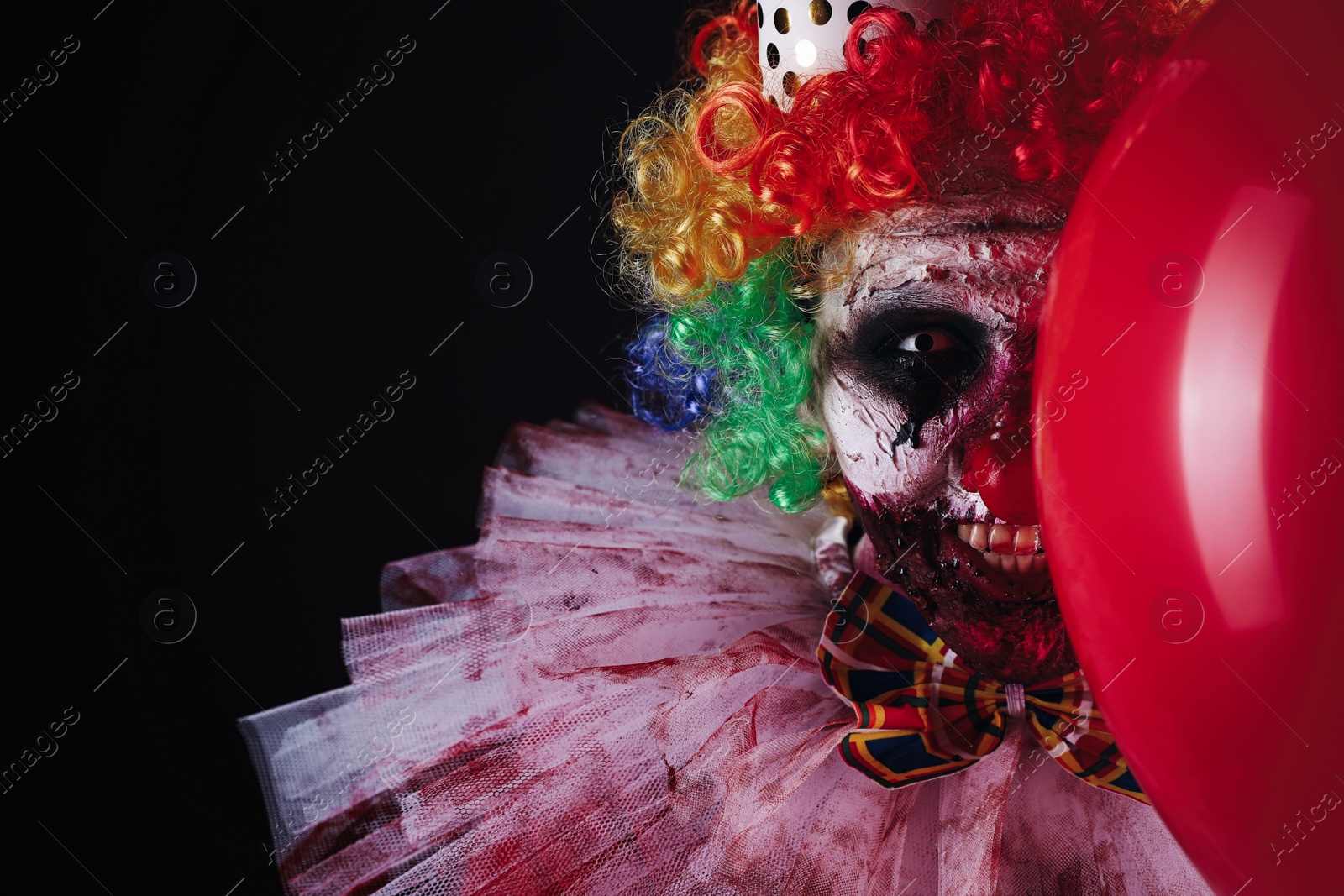 Photo of Terrifying clown with red air balloon on black background. Halloween party costume