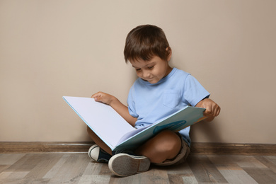 Cute little boy reading book on floor near beige wall