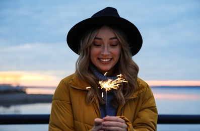 Woman in warm clothes holding burning sparkler near river