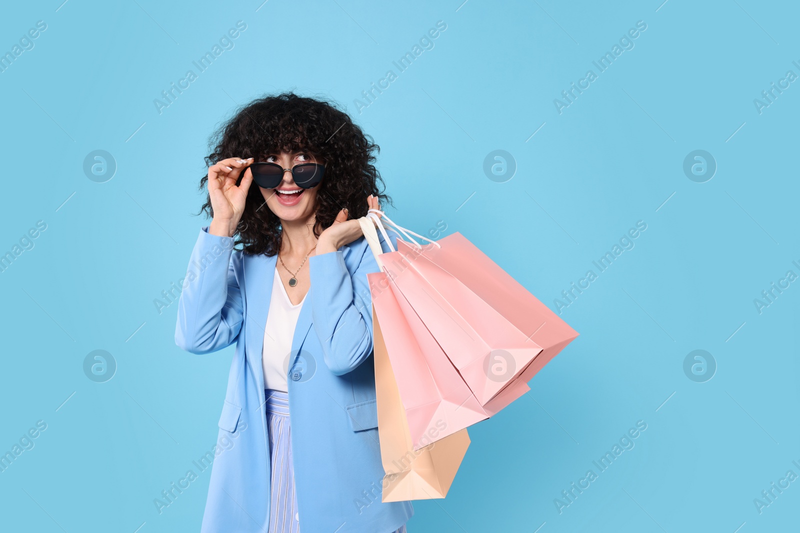 Photo of Happy young woman with shopping bags and stylish sunglasses on light blue background. Space for text