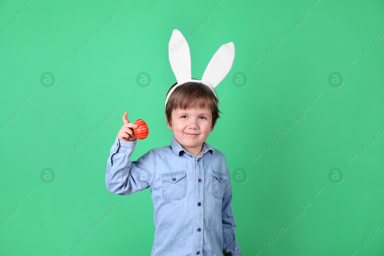 Photo of Cute little boy wearing bunny ears with dyed Easter egg on green background