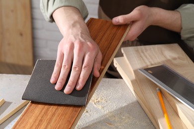 Man polishing wooden plank with sandpaper at grey table indoors, closeup