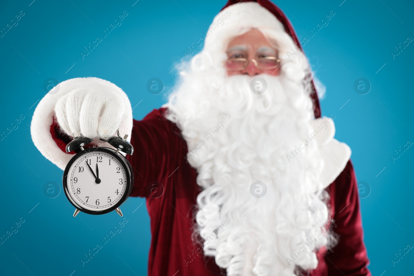 Photo of Santa Claus holding alarm clock on blue background, focus on hand. Christmas countdown