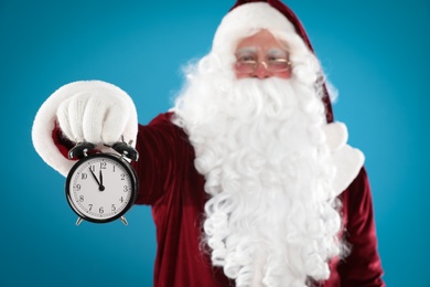 Photo of Santa Claus holding alarm clock on blue background, focus on hand. Christmas countdown