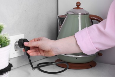 Photo of Woman plugging electric kettle into power socket at white table indoors, closeup