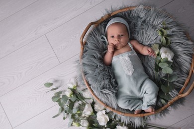 Photo of Adorable newborn baby lying in basket with faux fur and flowers on floor, top view. Space for text 