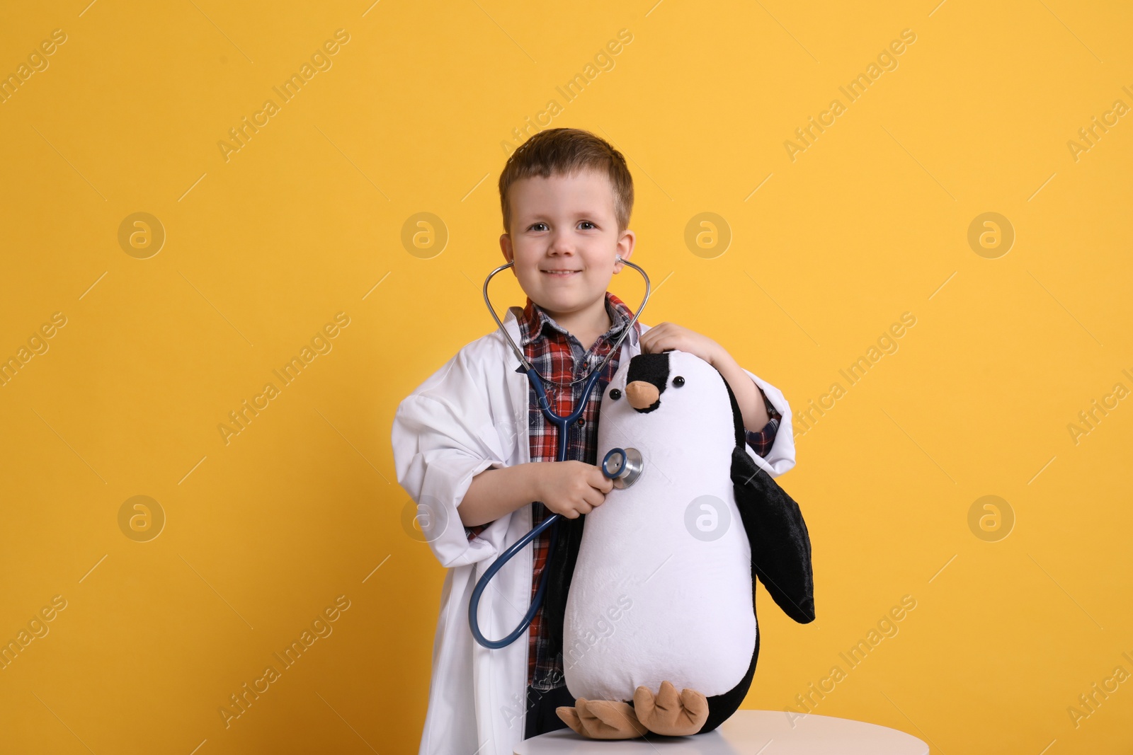 Photo of Cute little boy in pediatrician's uniform playing with stethoscope and toy penguin on yellow background