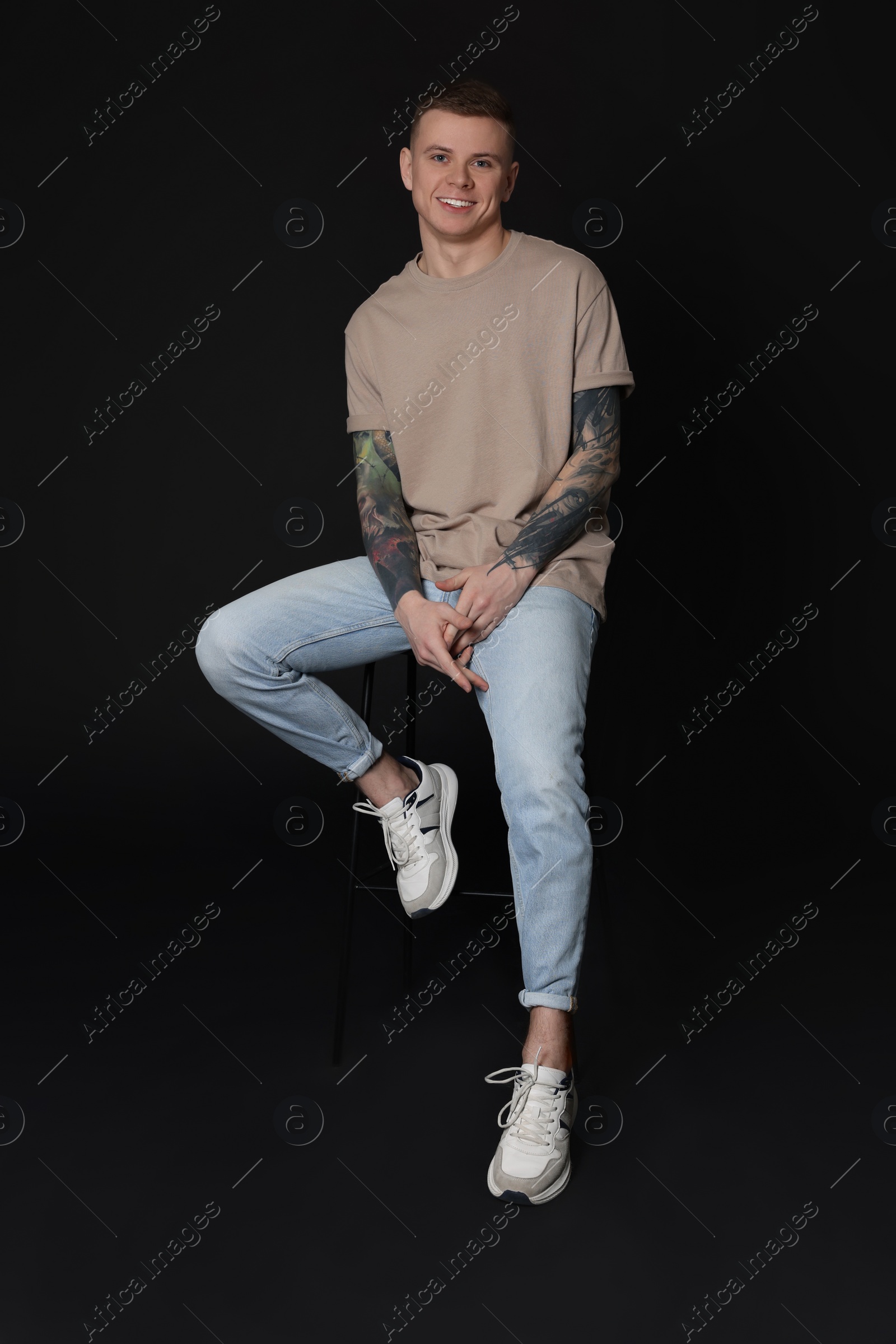 Photo of Smiling young man with tattoos sitting on stool against black background