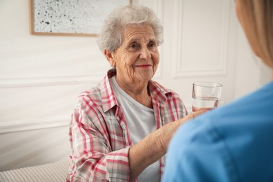 Photo of Young caregiver giving water to senior woman in room. Home care service