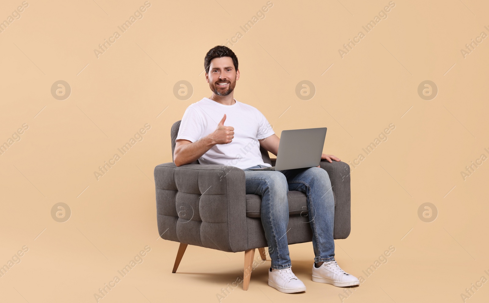 Photo of Happy man with laptop sitting in armchair and showing thumb up against beige background. Space for text
