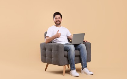 Photo of Happy man with laptop sitting in armchair and showing thumb up against beige background. Space for text