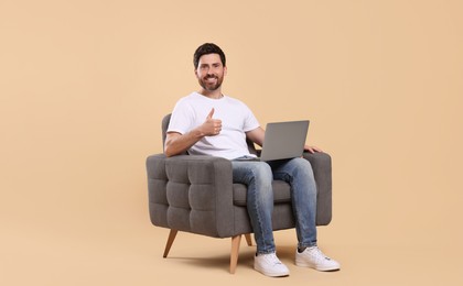 Happy man with laptop sitting in armchair and showing thumb up against beige background. Space for text