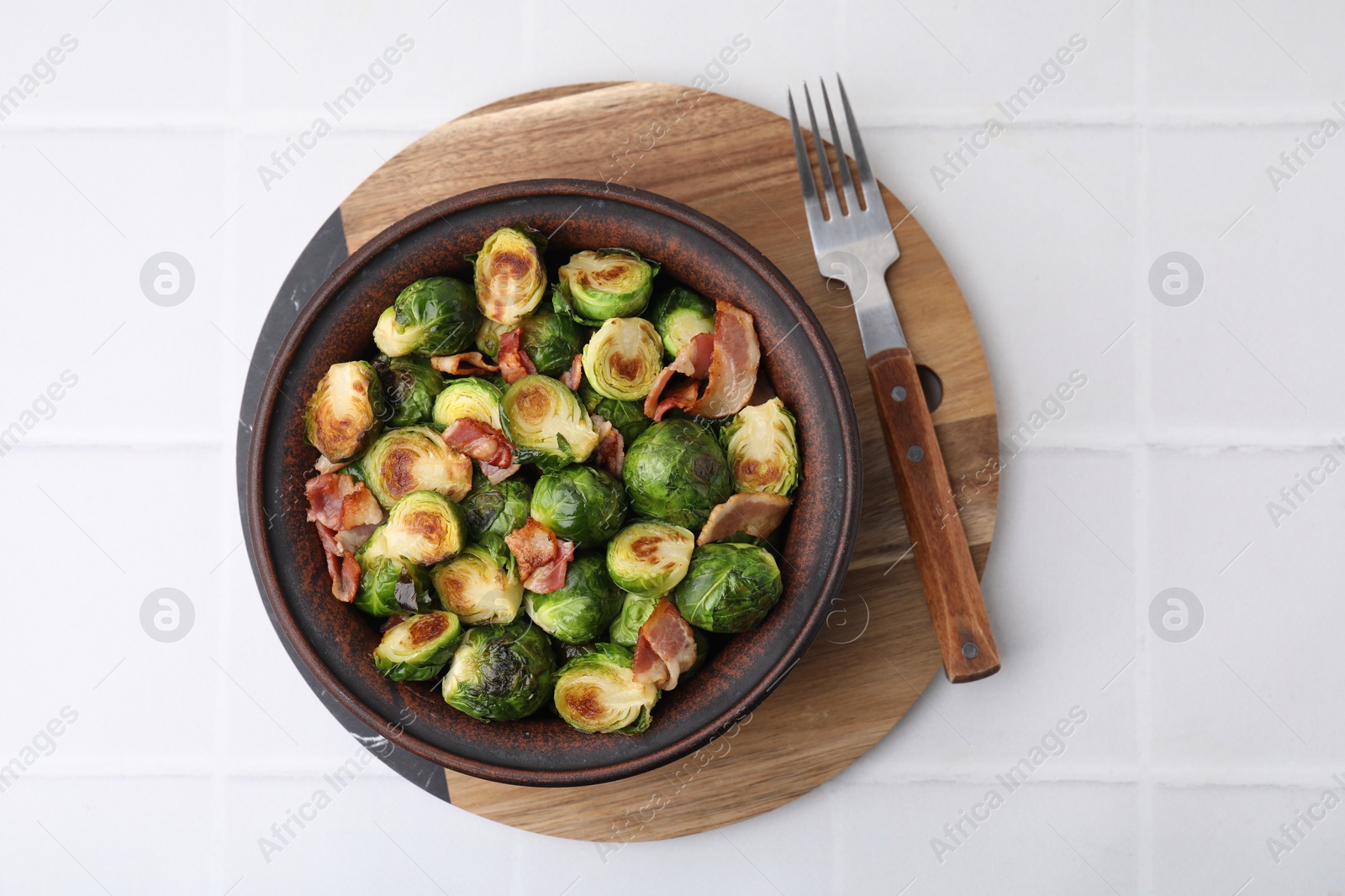 Photo of Delicious roasted Brussels sprouts and bacon in bowl on white tiled table, top view