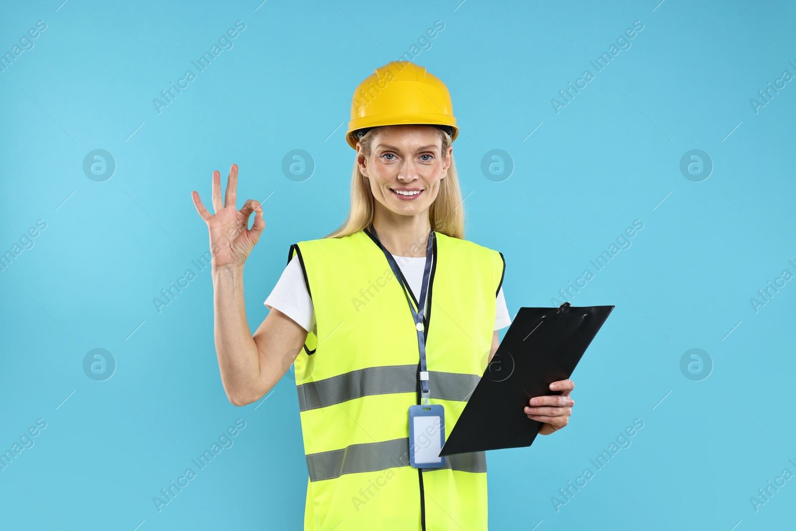 Photo of Engineer in hard hat holding clipboard and showing ok gesture on light blue background