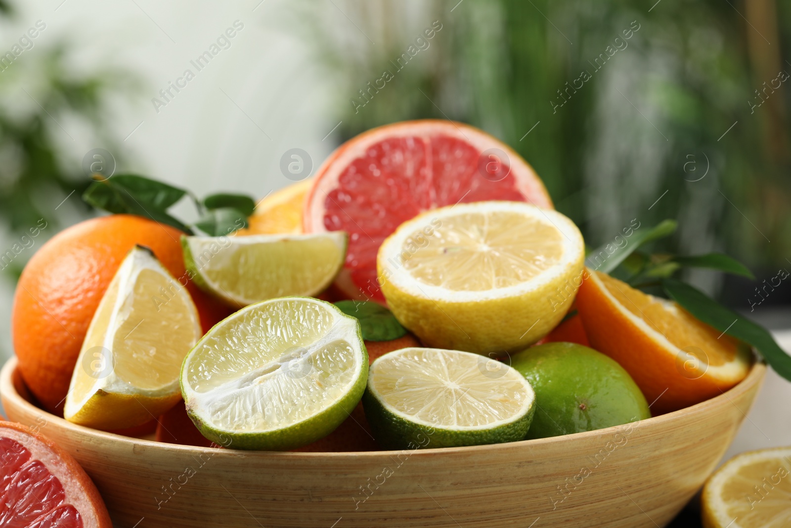 Photo of Different fresh citrus fruits and leaves in bowl against blurred background, closeup