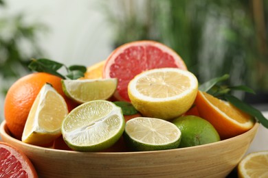 Photo of Different fresh citrus fruits and leaves in bowl against blurred background, closeup