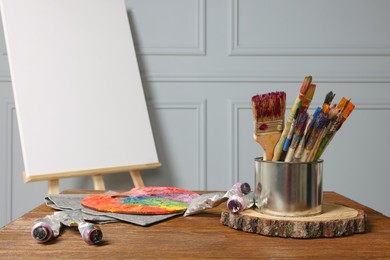 Easel with blank canvas and different art supplies on wooden table near grey wall