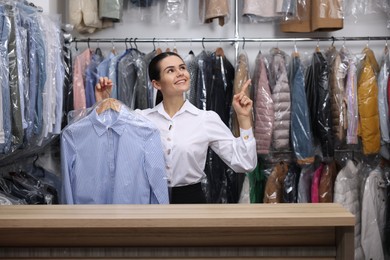 Photo of Dry-cleaning service. Happy worker holding hanger with shirt in plastic bag and pointing at something indoors, space for text