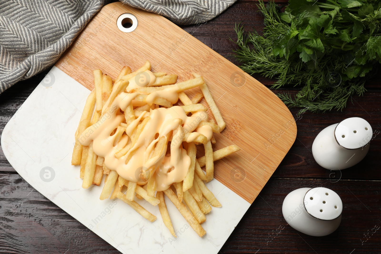 Photo of Delicious french fries with cheese sauce, greens and spice shakers on wooden table, flat lay