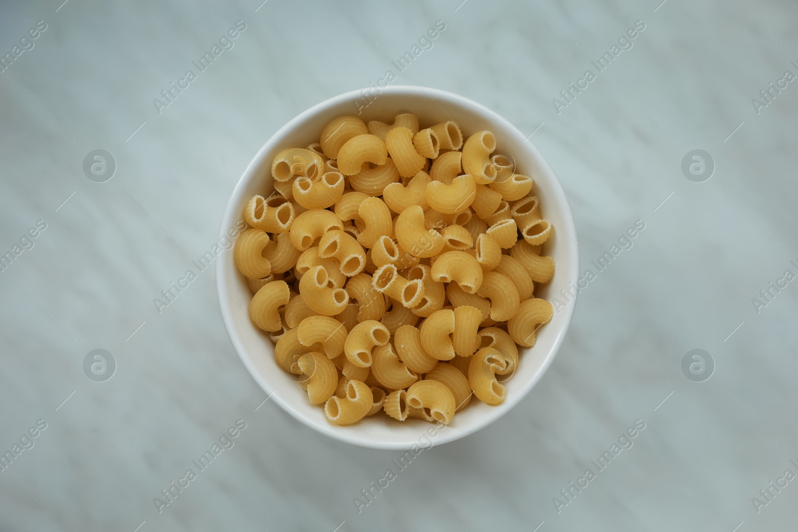 Photo of Bowl with uncooked horns pasta on white marble table, top view