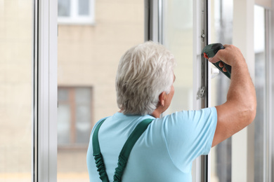 Mature construction worker repairing plastic window with electric screwdriver indoors