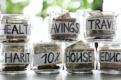 Photo of Glass jars with money for different needs on table against blurred background