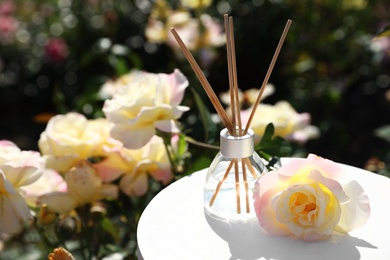 Photo of Reed air freshener with oil and fresh rose on table against blurred background. Space for text