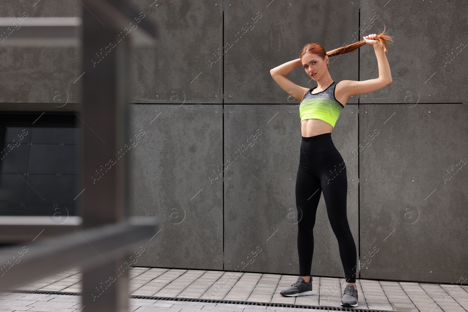 Photo of Beautiful woman in stylish gym clothes posing near grey wall on street