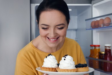 Photo of Happy overweight woman with cakes near fridge in kitchen
